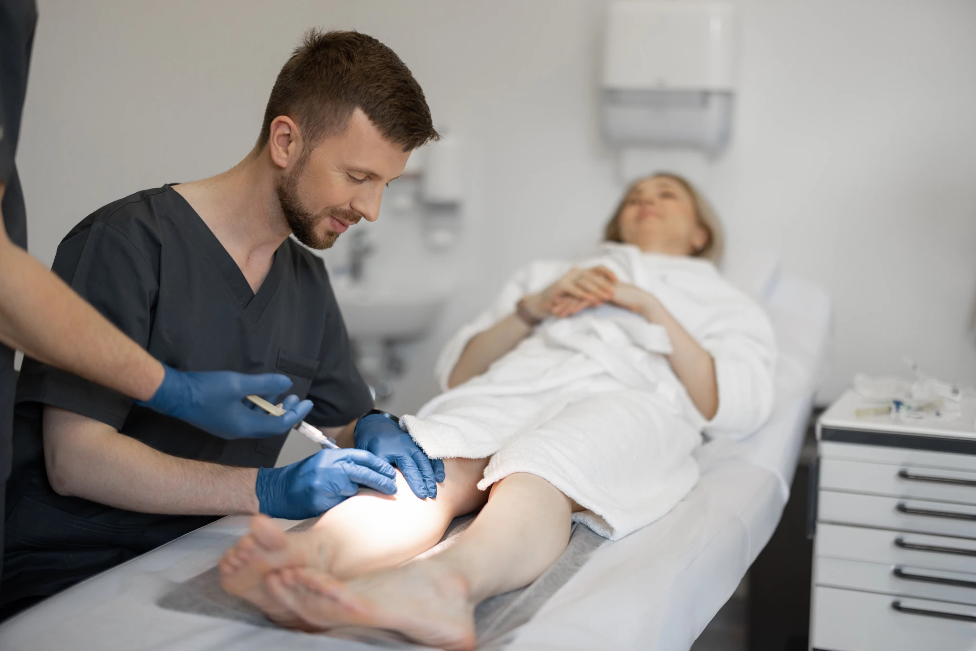 Médico aplica escleroterapia na perna de uma paciente deitada na maca